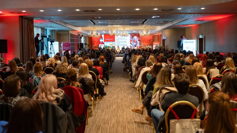 O mulțime de persoane stă așezată pe scaune într-o sală de conferințe, orientată spre o scenă cu un ecran mare și un podium iluminat.