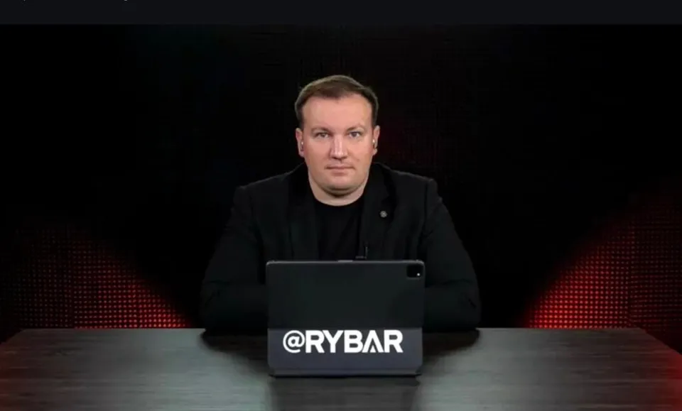 Un bărbat cu părul scurt și haine negre stă la o masă în fața unui ecran negru, având un dispozitiv cu textul @RYBAR în față.