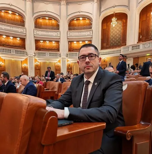 Un bărbat cu ochelari și cravată este așezat într-un scaun de piele într-o sală de conferințe cu balcoane decorate.
