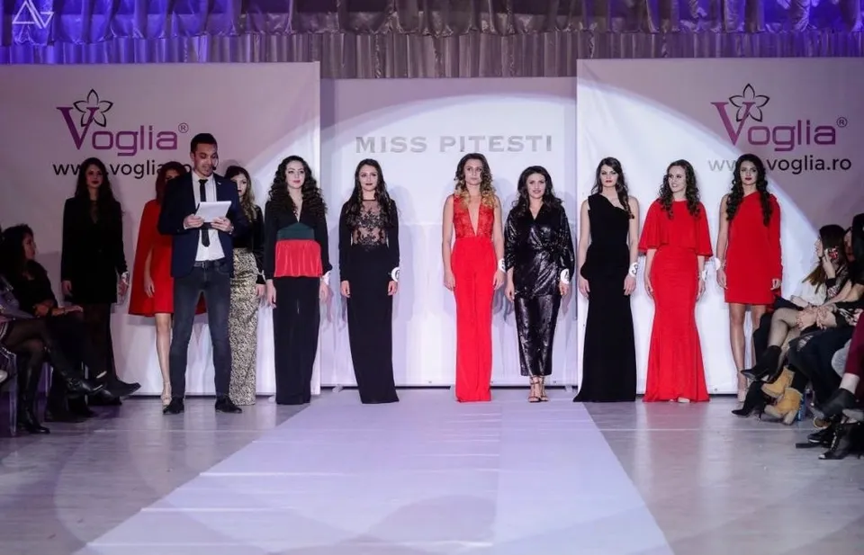 Oamenii stau pe un podium alb, în timp ce un grup de femei în rochii colorate pășește pe o pistă de catwalk.