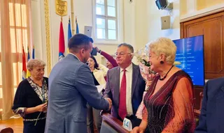 Un bărbat într-un costum albastru strânge mâna unui bărbat cu cravată roșie, în timp ce o femeie cu părul blond și o altă femeie privesc.