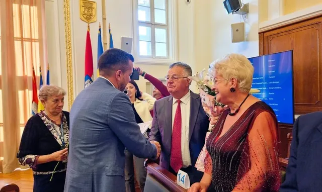Un bărbat într-un costum albastru strânge mâna unui bărbat cu cravată roșie, în timp ce o femeie cu părul blond și o altă femeie privesc.