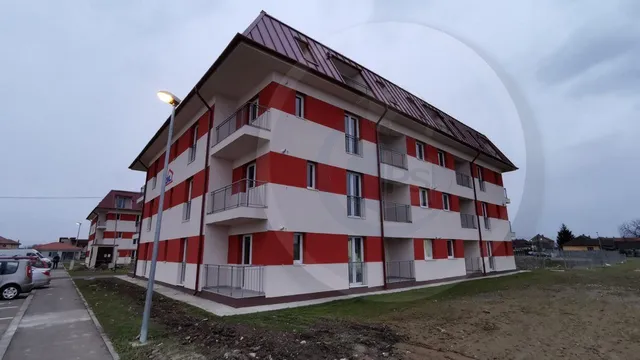 Un bloc de apartamente cu fațadă albă și roșie, dotat cu balcoane și acoperiș înclinat, este situat sub un cer înnorat.
