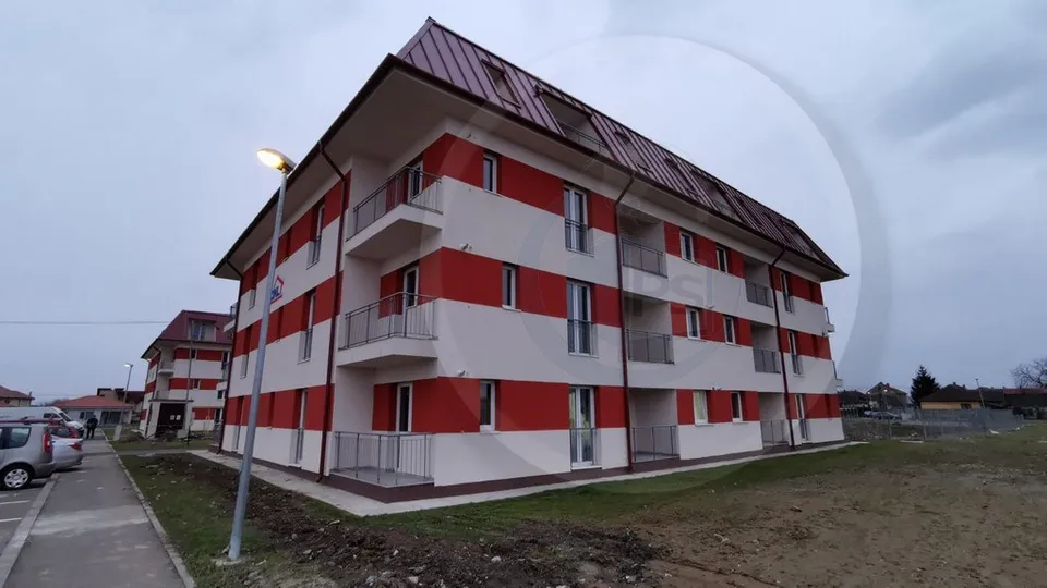Un bloc de apartamente cu fațadă albă și roșie, dotat cu balcoane și acoperiș înclinat, este situat sub un cer înnorat.