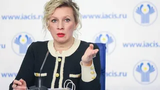 O femeie cu părul blond și roșu poartă o jachetă neagră cu detalii albe și butoane aurii, stând în fața unui fundal alb cu logouri albastre.