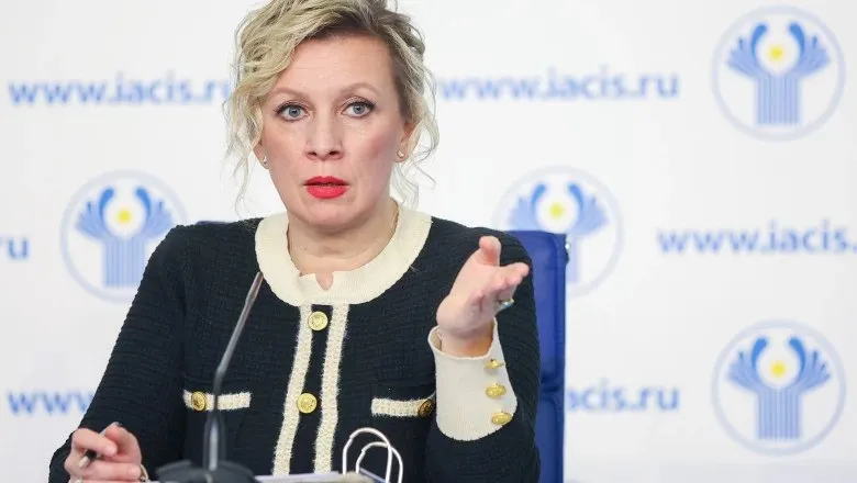 O femeie cu părul blond și roșu poartă o jachetă neagră cu detalii albe și butoane aurii, stând în fața unui fundal alb cu logouri albastre.