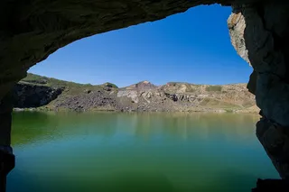 Vederea unui lac de culoare turcoaz prin deschiderea unei peșteri sau a unei cavități stâncoase spre un peisaj muntos cu versanți de piatră.