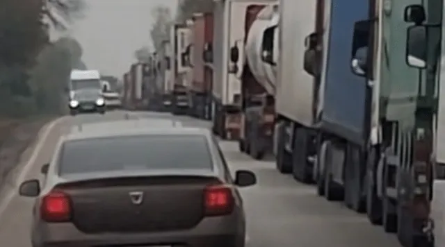 O linie de camioane mari este parcată pe marginea unui drum lângă o mașină de culoare închisă aflată în prim-plan.