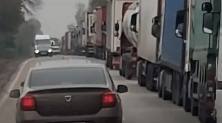 O linie de camioane mari este parcată pe marginea unui drum lângă o mașină de culoare închisă aflată în prim-plan.