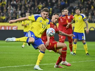 Un jucător în echipament galben și albastru lovește o minge de fotbal pe un teren verde în fața unor jucători în echipament roșu și mulțime.