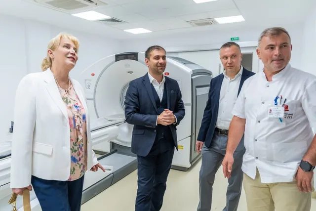 Patru persoane stau într-o încăpere medicală lângă un aparat de scanare mare, într-un spațiu cu pereți albi și lumină artificială.