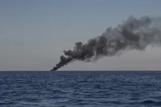 O coloană de fum negru se ridică deasupra orizontului deasupra unei suprafețe de apă de culoare albastră sub un cer senin.
