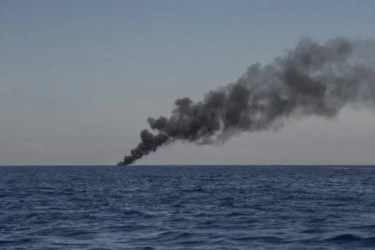 O coloană de fum negru se ridică deasupra orizontului deasupra unei suprafețe de apă de culoare albastră sub un cer senin.
