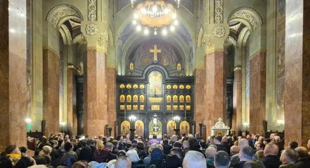 O mulțime de oameni stă în picioare în interiorul unei catedrale cu coloane mari, sub un candelabru și în fața unui iconostas decorat cu icoane.