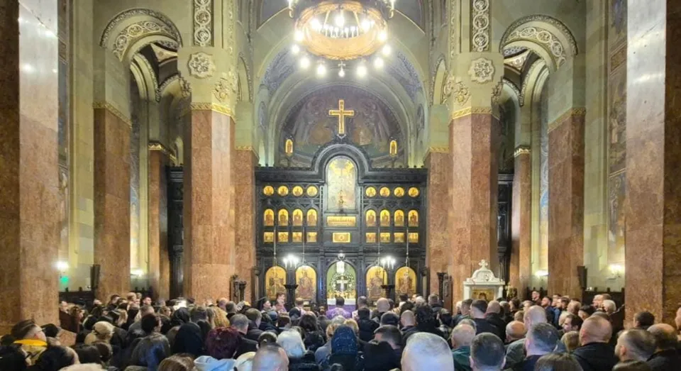 O mulțime de oameni stă în picioare în interiorul unei catedrale cu coloane mari, sub un candelabru și în fața unui iconostas decorat cu icoane.