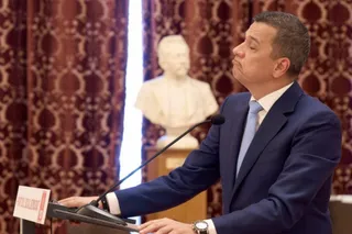 Un bărbat într-un costum albastru și cravată deschisă stă în fața unui microfon, cu o bustă albă în fundalul neclar.
