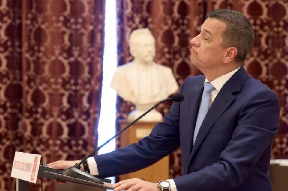 Un bărbat într-un costum albastru și cravată deschisă stă în fața unui microfon, cu o bustă albă în fundalul neclar.