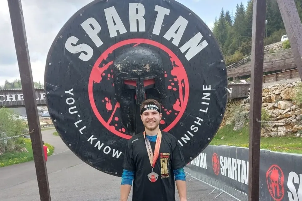 Un bărbat cu medalie la gât stă în fața unui scut mare pe care scrie SPARTAN și YOU'LL KNOW WHEN YOU CROSS THE FINISH LINE.
