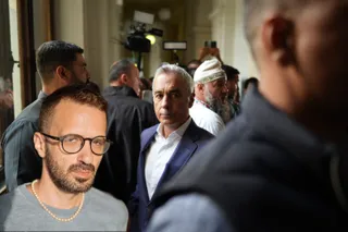 Un grup de bărbați stă strâns într-un spațiu interior, un bărbat cu ochelari și colier de perle fiind în prim-plan, în timp ce alții privesc spre cameră.