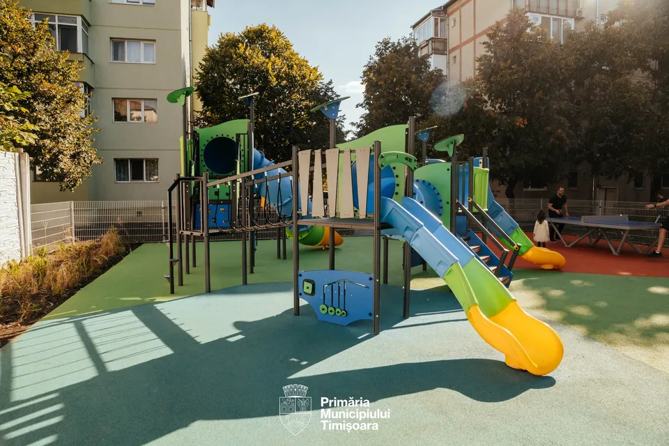 Un loc de joacă colorat cu tobogane, structuri de escaladare și o masă de tenis de masă este situat pe un suprafață de cauciuc verde și roșie.