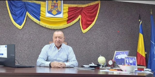 Un bărbat cu cămașă în dungi albe și albastre stă la un birou în fața unui drapaj tricolor cu un blazon.