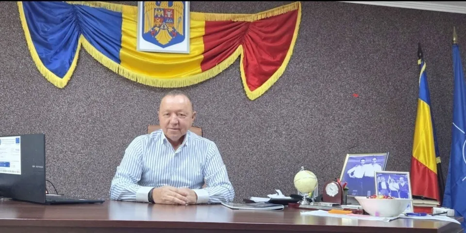 Un bărbat cu cămașă în dungi albe și albastre stă la un birou în fața unui drapaj tricolor cu un blazon.
