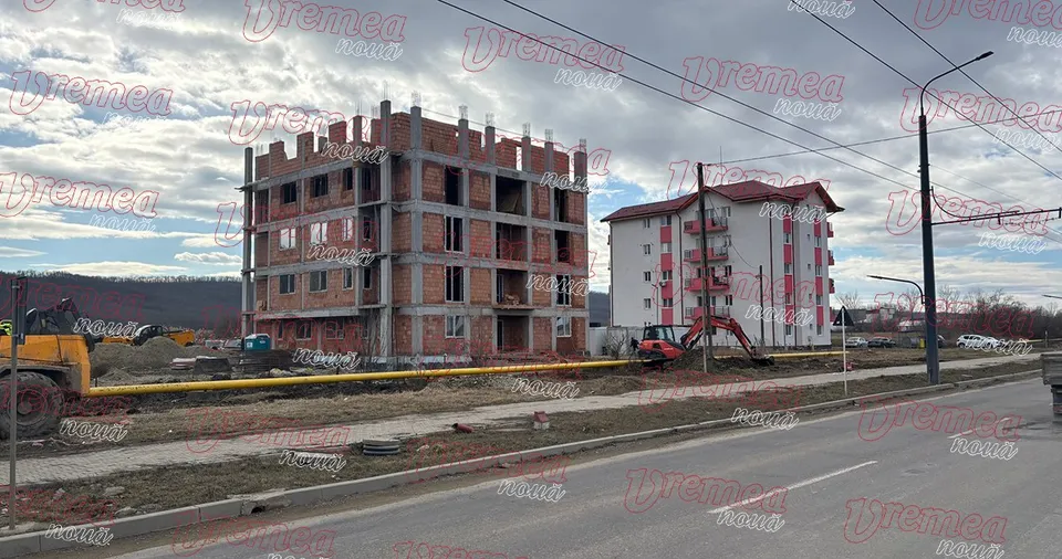 Un bloc de apartamente în construcție din cărămidă roșie stă lângă un bloc alb cu ferestre și balcoane sub un cer înnorat.