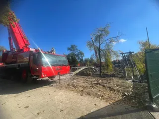 Un camion roșu cu o macara ridicată stă pe un teren cu pământ și resturi lângă o structură metală curbată sub un cer senin.