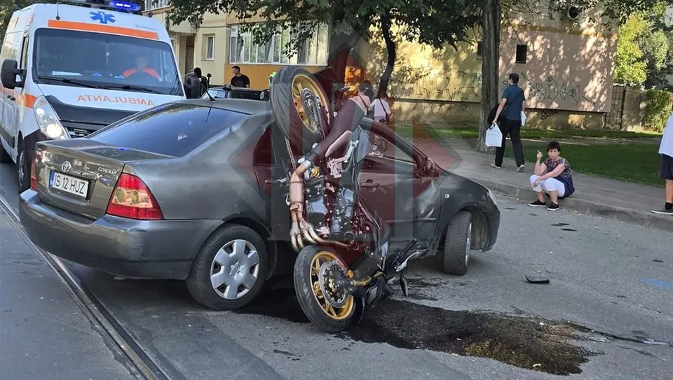 Un autoturism gri este răsturnat pe o motocicletă, în timp ce o ambulanță albă este parcată în planul secund.
