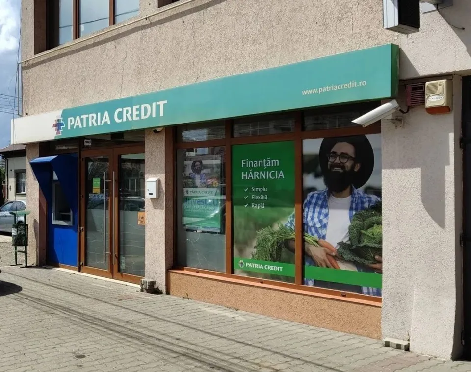 O clădire cu o vitrină ce afișează un panou verde cu textul Patria Credit și un poster cu un bărbat ținând legume verzi.
