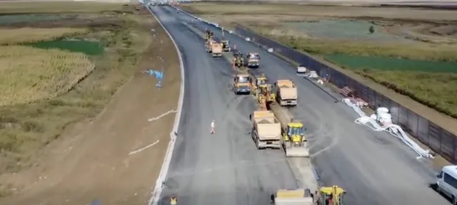 O vedere aeriană prezintă mai multe utilaje de construcții și camioane parcate pe o porțiune de drum asfaltat în curs de execuție, lângă un teren agricol.