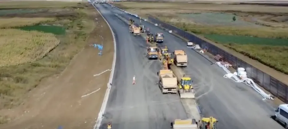 O vedere aeriană prezintă mai multe utilaje de construcții și camioane parcate pe o porțiune de drum asfaltat în curs de execuție, lângă un teren agricol.