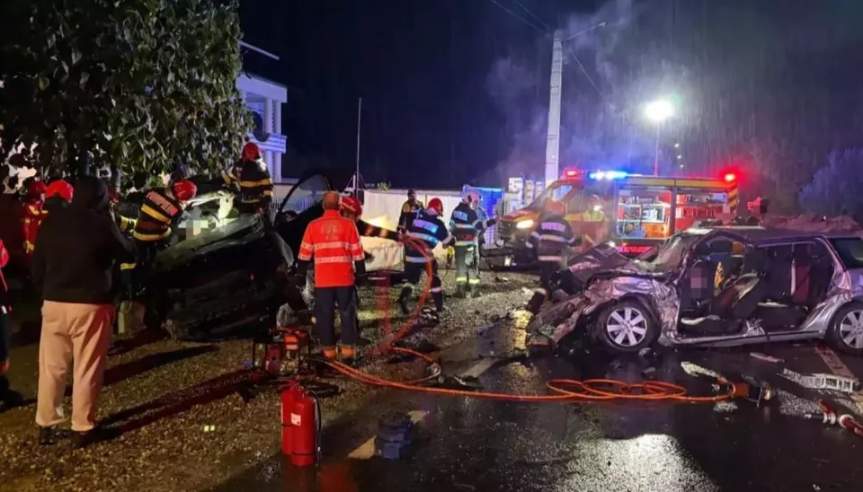 Echipe de intervenție și persoane în haine reflectorizante stau lângă două autoturisme distruse pe un drum umed, noaptea, lângă un vehicul de serviciu cu lumină albastră.