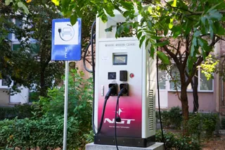 O stație de reîncărcare albă cu detalii roz și un semn albastru cu un simbol de vehicul electric sunt amplasate lângă vegetație.