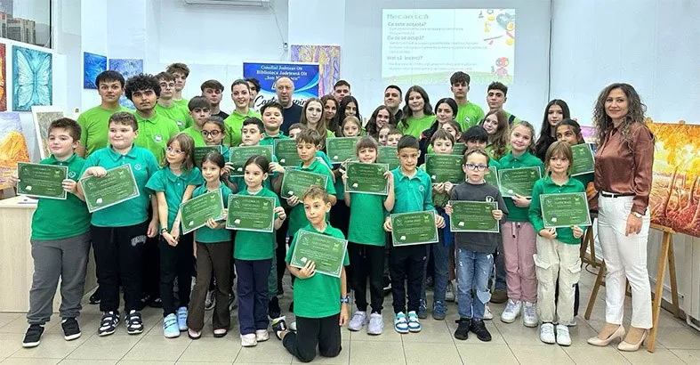 Un grup de copii și adulți stau în picioare într-o încăpere, mulți dintre ei ținând diplome verzi cu margini aurii.