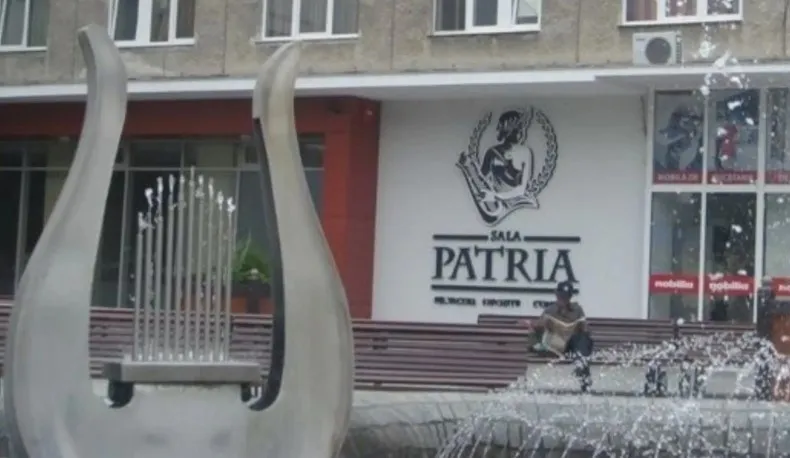 O sculptură metalică în formă de harpă se află în prim-plan, în fața unei clădiri cu inscripția Sala Patria.