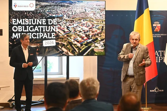 Un bărbat într-un sacou bej vorbește la un microfon în fața unui ecran mare și a unui steag, în timp ce un alt bărbat stă în picioare.
