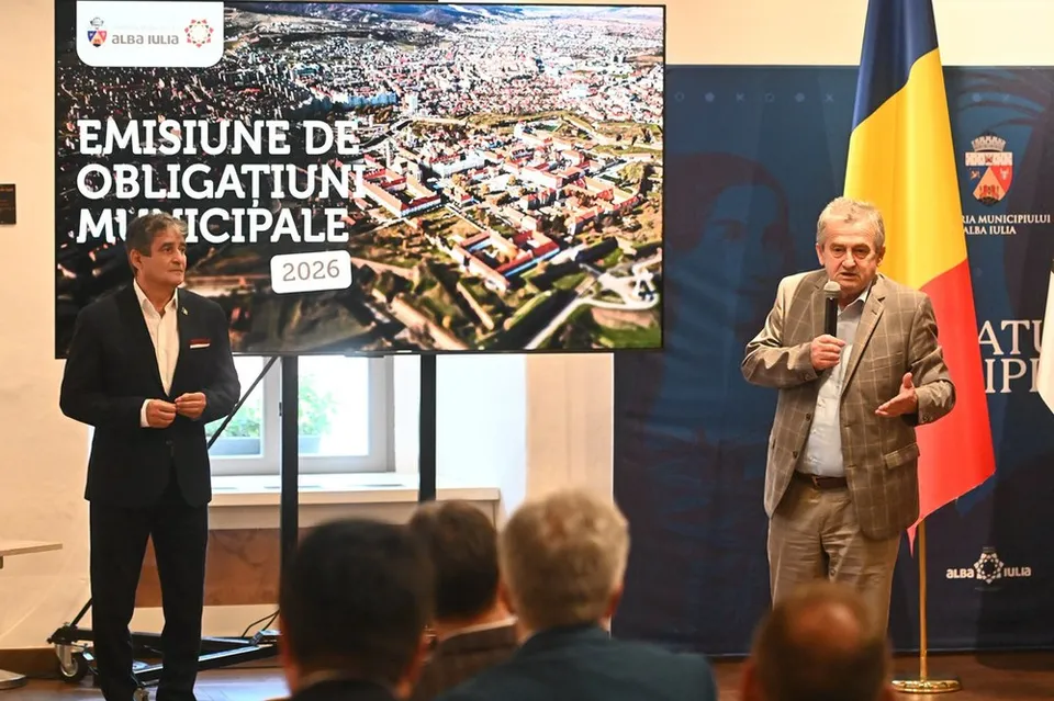 Un bărbat într-un sacou bej vorbește la un microfon în fața unui ecran mare și a unui steag, în timp ce un alt bărbat stă în picioare.