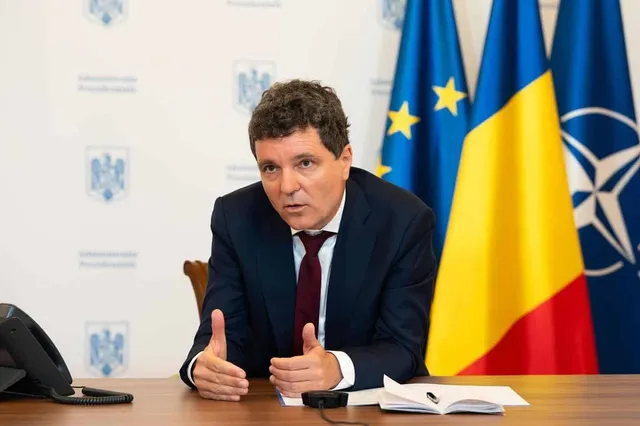 Un bărbat cu părul închis la culoare, purtând sacou închis și cravată roșie, stă la o masă de lemn în fața unor steaguri.