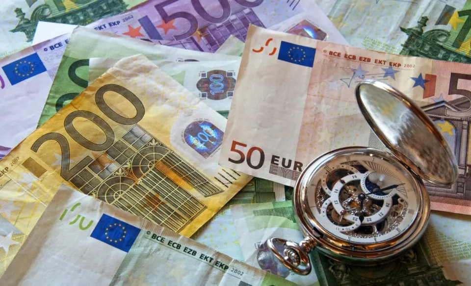 Un ceas de buzunar argintiu cu mecanism expus este așezat pe mai multe bancnote de euro împrăștiate.
