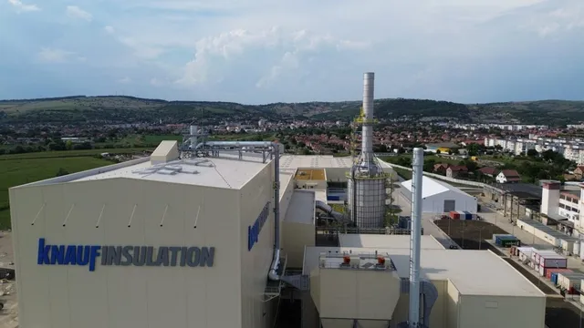 O vedere aeriană a unei clădiri industriale de culoare deschisă cu logoul Knauf Insulation, având o turută înaltă și un oraș în fundal.