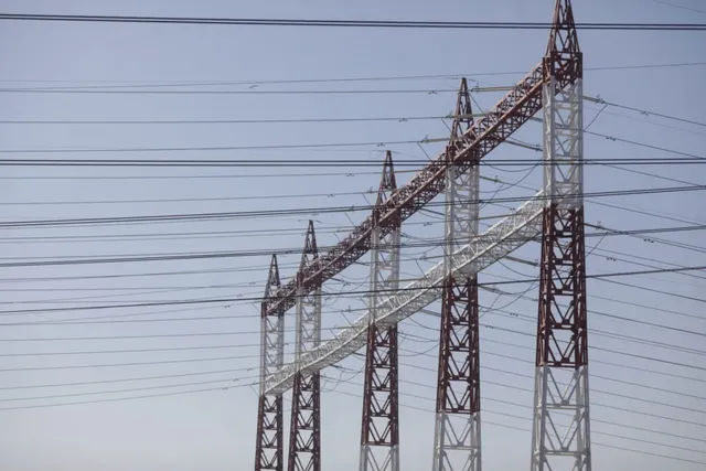 Mai multe turnuri metalice de înaltă tensiune susțin numeroase cabluri electrice întinse pe un cer deschis.