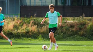 Un tânăr jucător de fotbal într-un echipament verde și negru conduce un balon pe un teren de gazon verde.