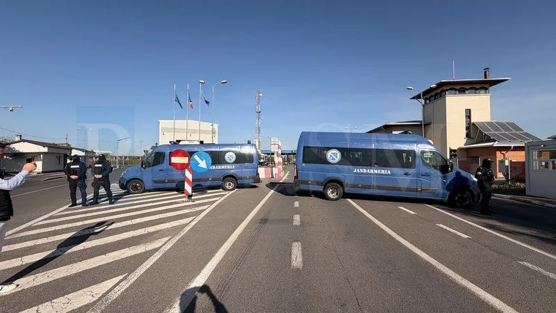 Două microbuze albastre cu inscripția Jandarmeria sunt parcate pe un drum asfaltat lângă clădiri și un turn de observație sub un cer senin.