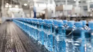 Rânduri de sticle de plastic cu lichid transparent și capace albastre sunt așezate pe o bandă de transport într-o unitate de producție.