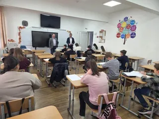 Elevi stau la bănci într-o sală de clasă, în timp ce doi bărbați stau în picioare în fața unui ecran de monitor.