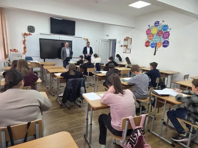 Elevi stau la bănci într-o sală de clasă, în timp ce doi bărbați stau în picioare în fața unui ecran de monitor.