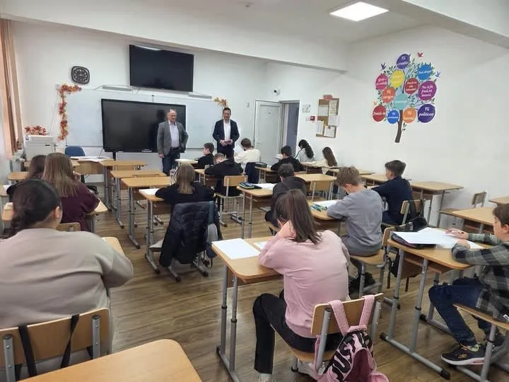 Elevi stau la bănci într-o sală de clasă, în timp ce doi bărbați stau în picioare în fața unui ecran de monitor.