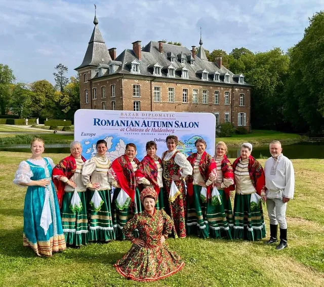 O grupă de persoane poartă costume tradiționale colorate și stă în fața unui panou mare și a unei clădiri mari cu acoperiș înclinat pe un prăjlin.