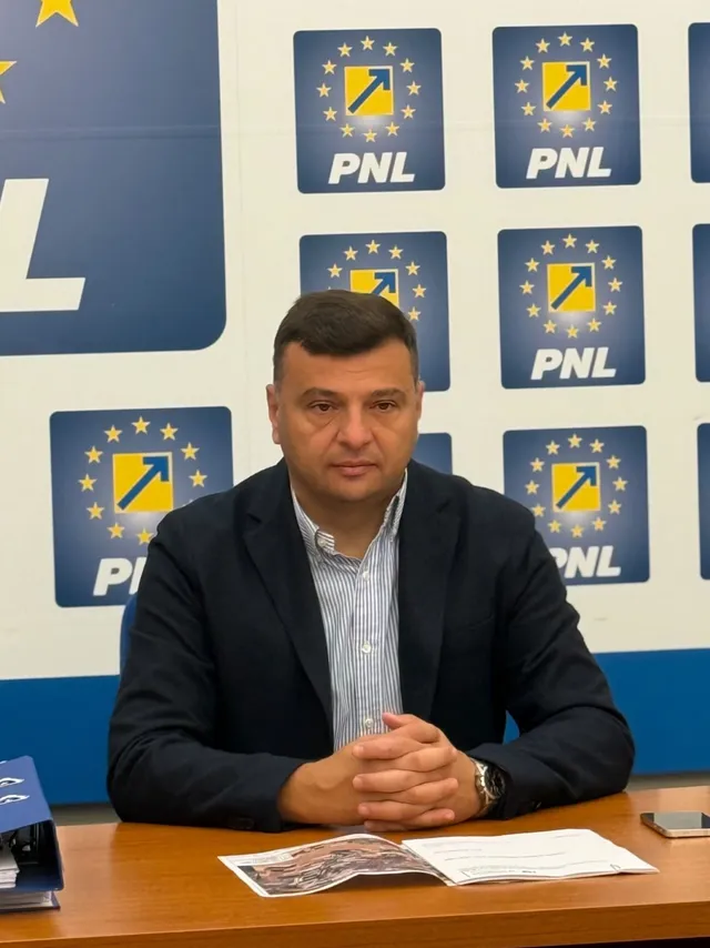 Un bărbat cu sacou închis și cămașă cu dungi verticale stă la o masă cu documente deschise în fața unui panou cu logo-uri repetitive.
